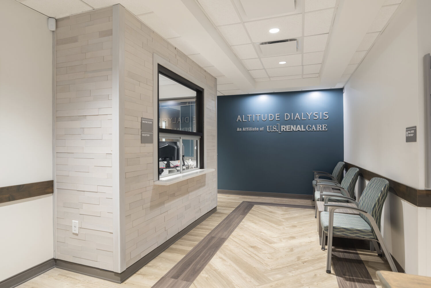 Reception window with a light stone accent wall, blue feature wall with Altitude Dialysis signage, patterned waiting chairs, wood-look flooring, and overhead acoustic ceiling panels.