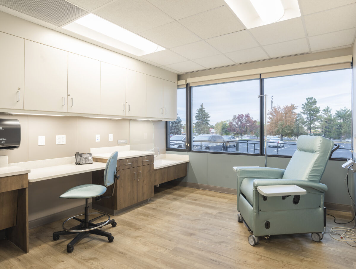 Private treatment room with a teal recliner, built-in work counter with storage cabinets, wall-mounted utilities, large windows with outdoor views, and wood-look flooring.