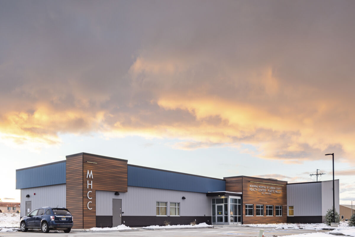 Exterior of the health center at sunset with wood and metal façades, MHCC signage on the corner tower, large windows, a parked vehicle, and snow-covered landscaping under a colorful sky.