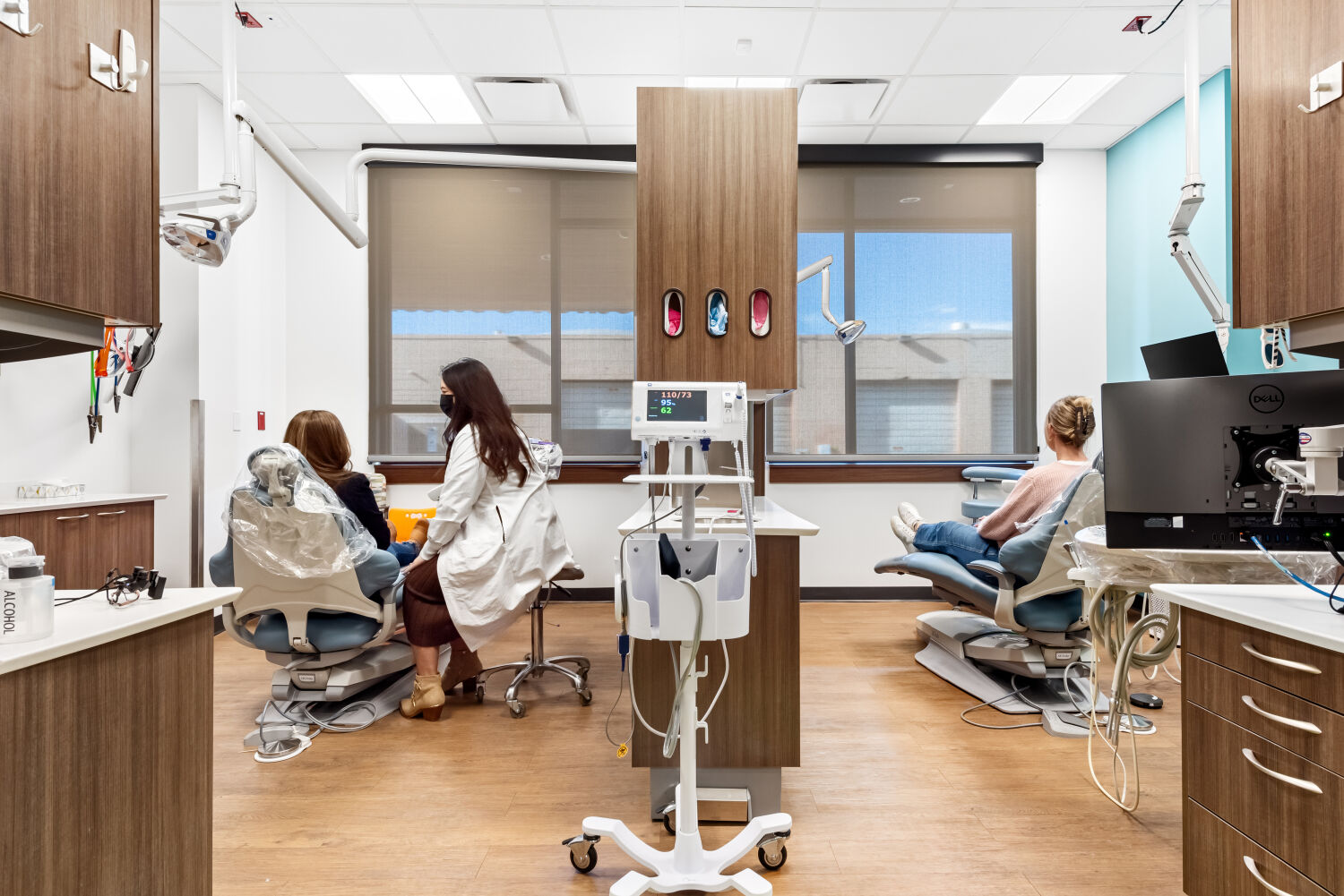 Dual dental operatory with two treatment chairs, overhead exam lights, wood cabinetry, clinical equipment on a mobile cart, and large windows with roller shades.