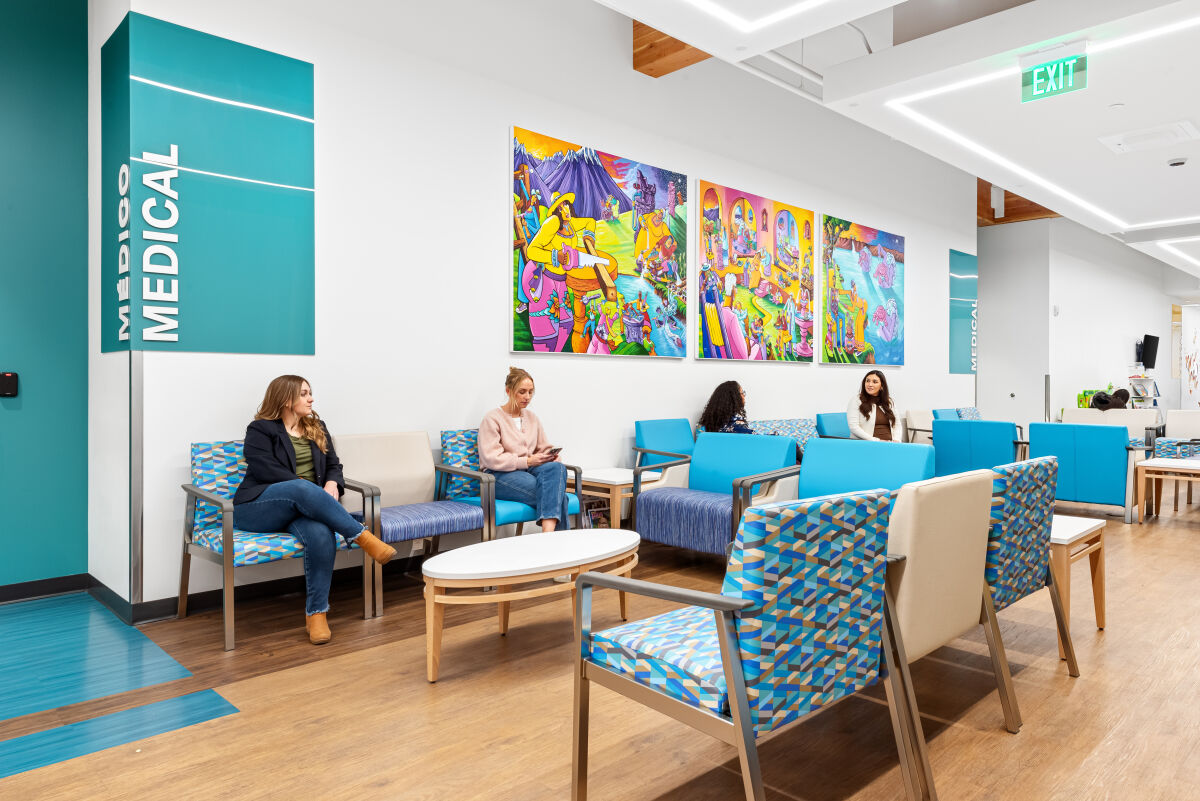 Bright waiting area with turquoise seating, wood flooring, and three large colorful murals on the wall beneath a ceiling with linear lighting; “MEDICAL” sign visible at left.