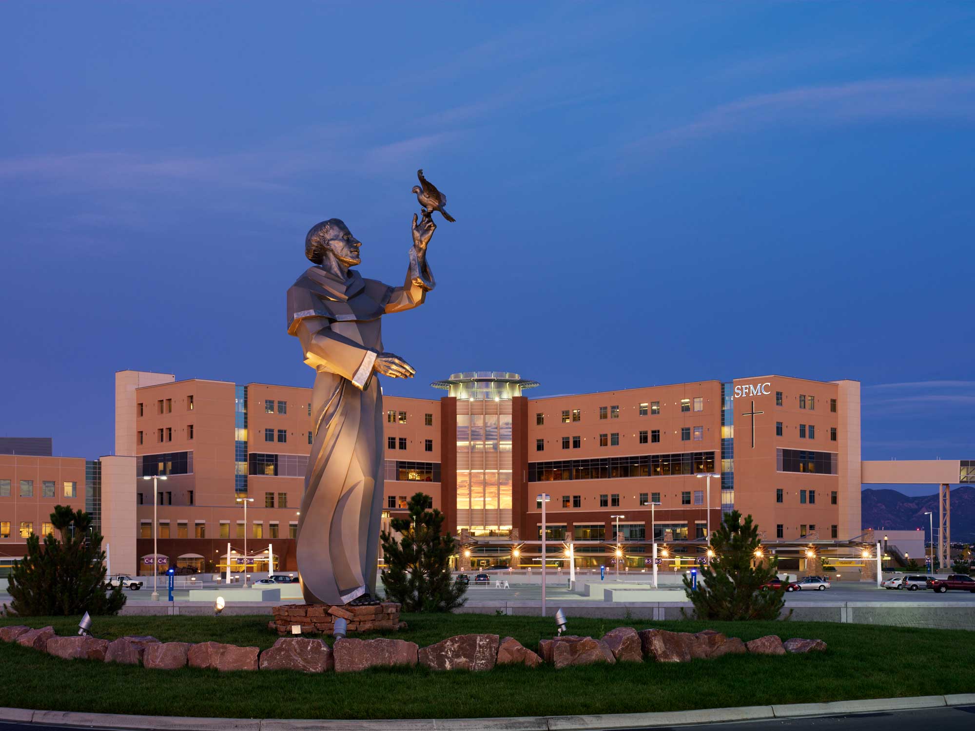 Exterior view of a hospital building at night, illuminated by soft, warm lighting. A prominent statue of St. Francis is featured in the foreground, surrounded by well-manicured landscaping and reflecting a serene ambiance.