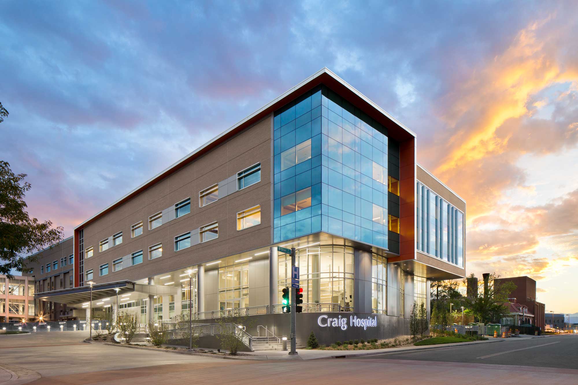 Nighttime exterior view of Craig Hospital, featuring a modern design with large glass windows and warm interior lighting. The building is surrounded by landscaped greenery, creating a welcoming and sophisticated appearance.