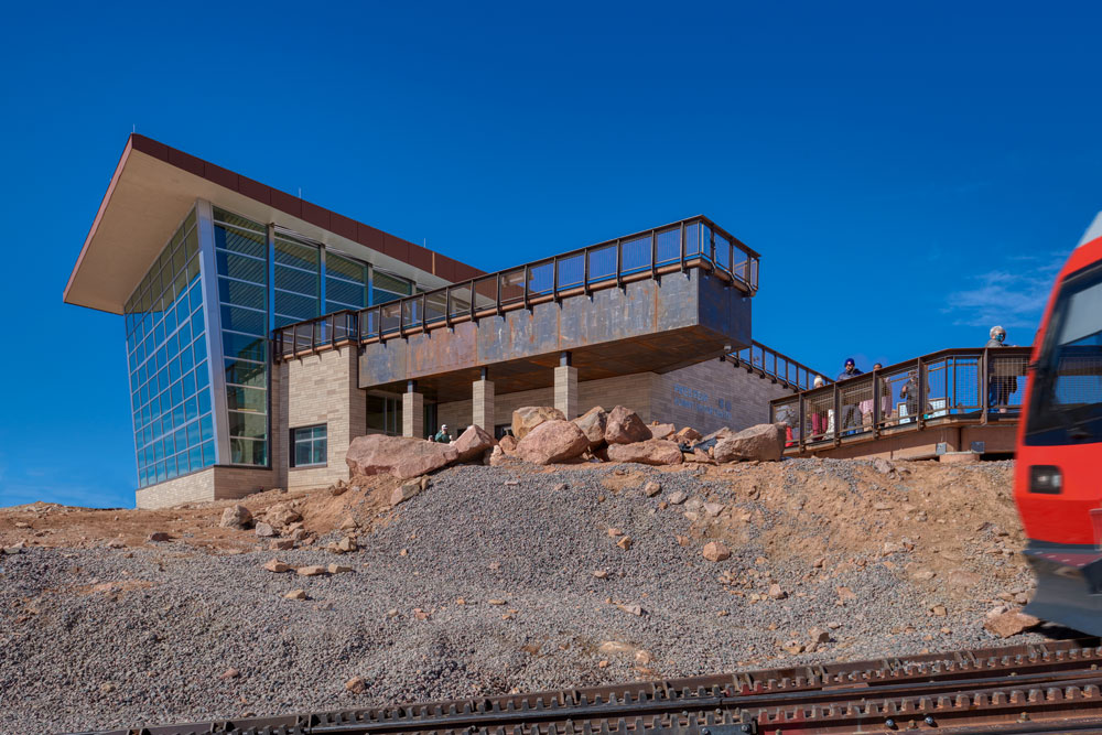 Aerial view of the Pikes Peak Visitor Center and Cog Railway, showcasing the modern summit complex with a sleek, low-profile design that blends into the mountain landscape. The railway tracks lead to the facility, offering visitors access to stunning panoramic views.